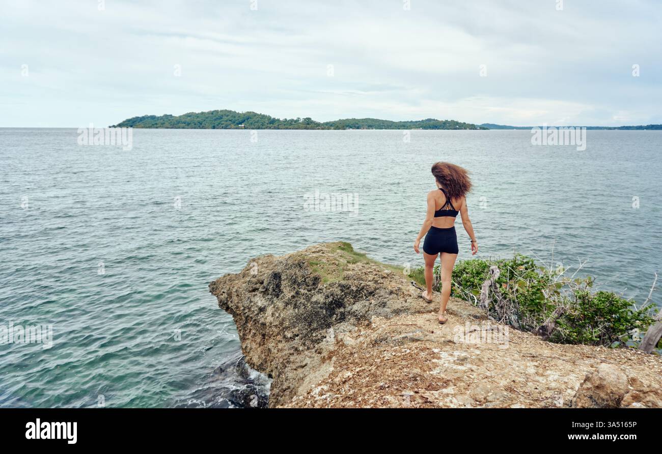 La donna ispanica con i capelli ricci cammina lungo una spiaggia illuminata dal sole. Un'immagine di viaggio rilassata e di stile di vita con paesaggi costieri e ampio spazio per il branding. Ideale per campagne di vacanza al mare, blog sul mare e promozioni estive. Foto Stock
