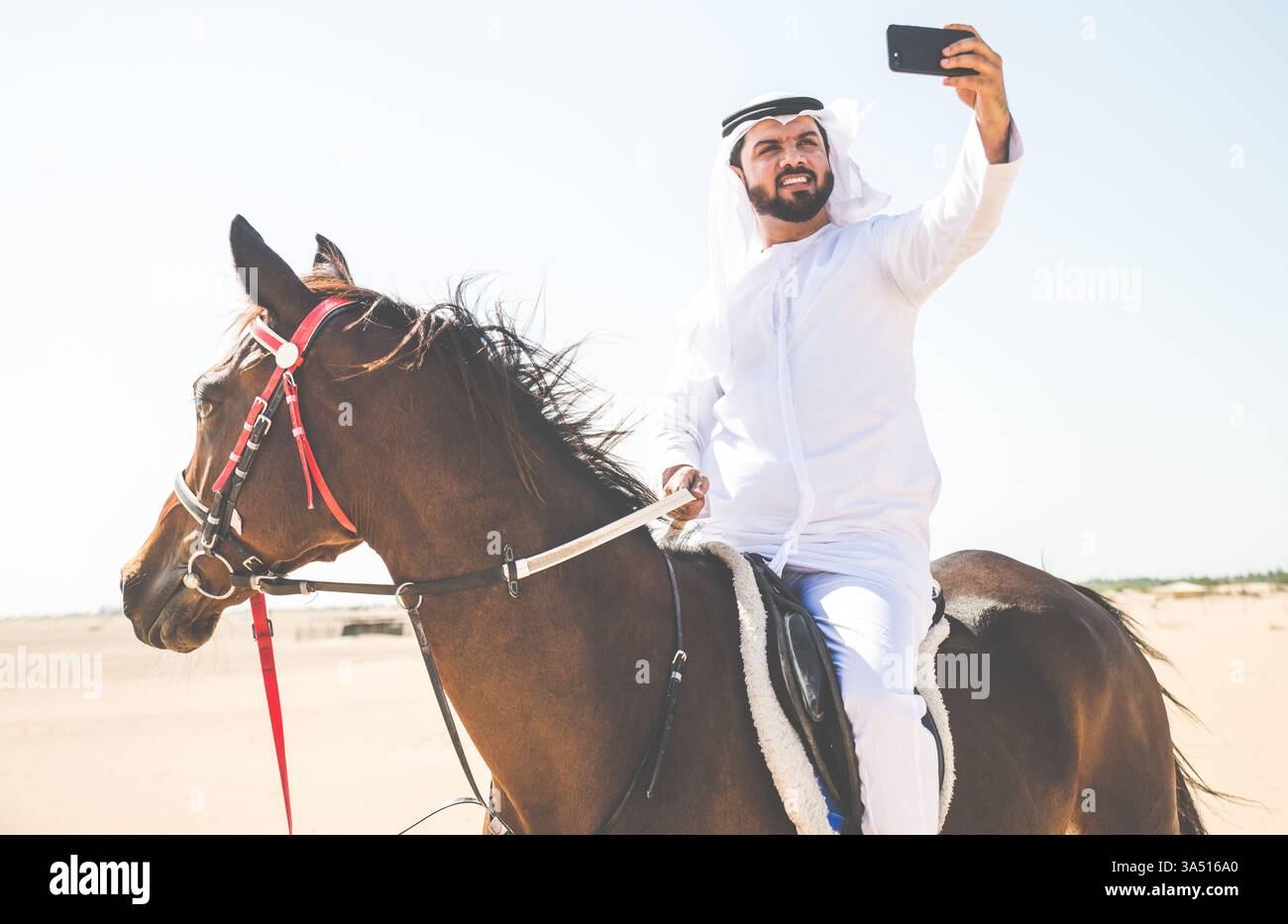 Un giovane che indossa Kandura cavalca un cavallo attraverso un paesaggio desertico negli Emirati. Questa scena di abbigliamento tradizionale mette in risalto il patrimonio mediorientale e la vita equestre in un suggestivo ambiente desertico. Ideale per viaggi, cultura e progetti lifestyle di lusso. Foto Stock
