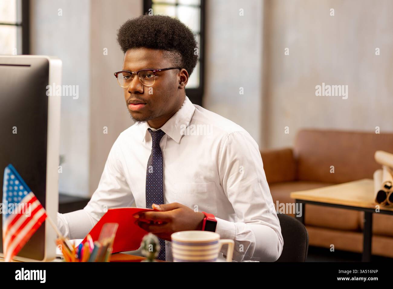 Un uomo nero serio seduto a un tavolo con un computer e un notebook in un ufficio moderno. Ritratto aziendale professionale adatto per la tecnologia, la finanza e le comunicazioni sul luogo di lavoro. Ideale per materiali aziendali, lavori digitali e immagini di leadership. Foto Stock