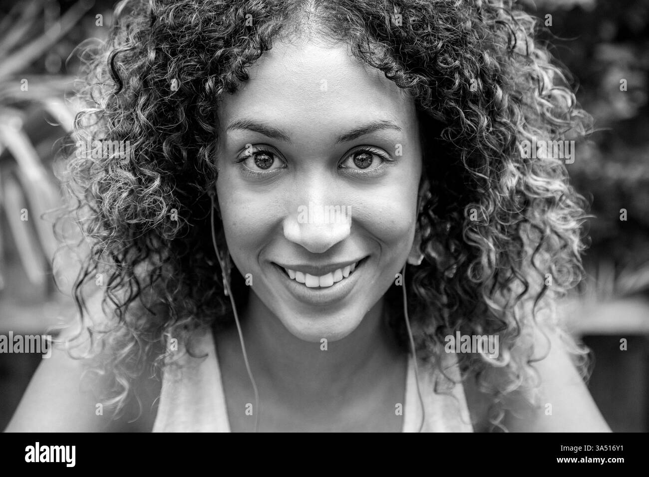 Una donna afroamericana sorridente con i capelli ricci che indossa gli auricolari siede su una panchina del parco. Sembra rilassata in un ambiente all'aperto, creando un ritratto di stile amichevole. Adatto a temi di benessere, svago all'aperto e stile di vita urbano. Foto Stock