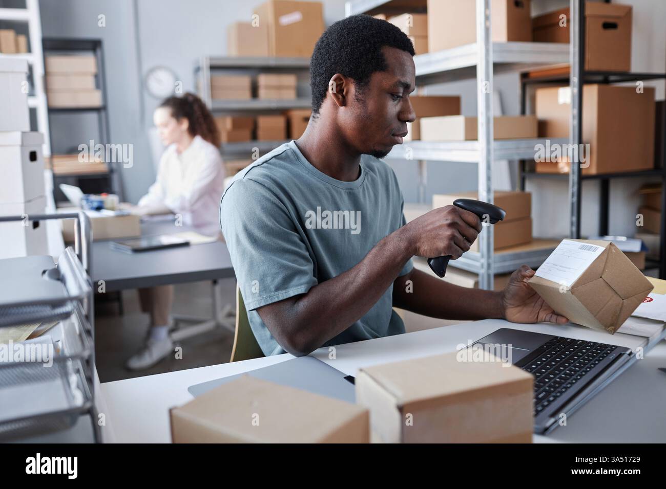 Un lavoratore di sesso maschile nero esegue la scansione di una scatola mentre è seduto a un tavolo vicino a una collega caucasica in un luogo di lavoro occupato. La scena suggerisce operazioni di logistica, imballaggio e magazzino, con un lavoro di squadra diversificato in azione. Perfetto per la grafica delle operazioni di e-commerce, delivery e punto vendita. Foto Stock
