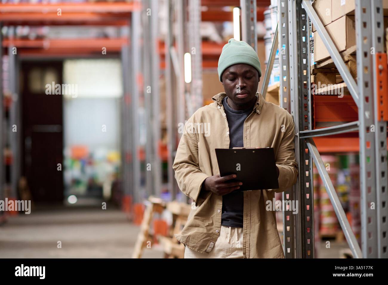 Un serio operaio di magazzino afroamericano esamina un programma di spedizione su un appunti, evidenziando la logistica in azione. Ideale per operazioni di magazzino, controllo dell'inventario e distribuzione di merci. Un'immagine di stock professionale adatta per argomenti di logistica, supply chain e distribuzione. Foto Stock