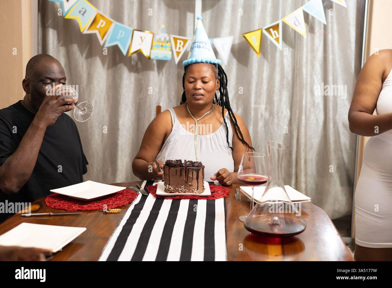 Il padre felice Nero sorride mentre la madre fetta una torta durante una festa di compleanno a casa. Questa calda festa al coperto offre amore per la famiglia, torta e insieme intorno al tavolo da pranzo. Un'immagine versatile per la vita familiare, le celebrazioni e le campagne di lifestyle. Foto Stock
