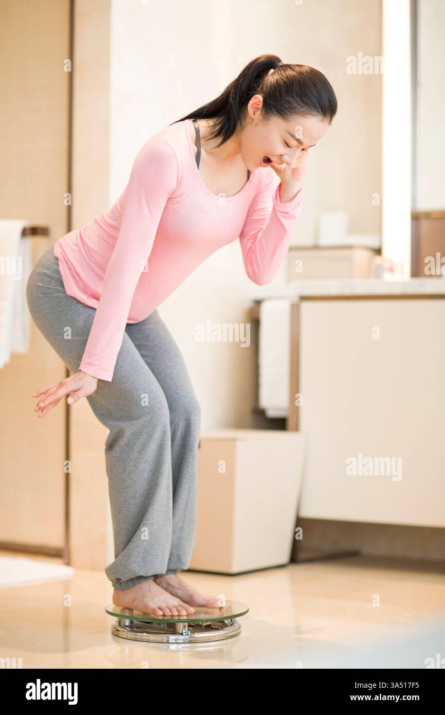 Giovane donna cinese che si pesa su una bilancia da bagno. Un momento sincero che mette in evidenza il monitoraggio del peso, la dieta e il benessere personale. Ideale per campagne di benessere, fitness e cura di sé. Foto Stock