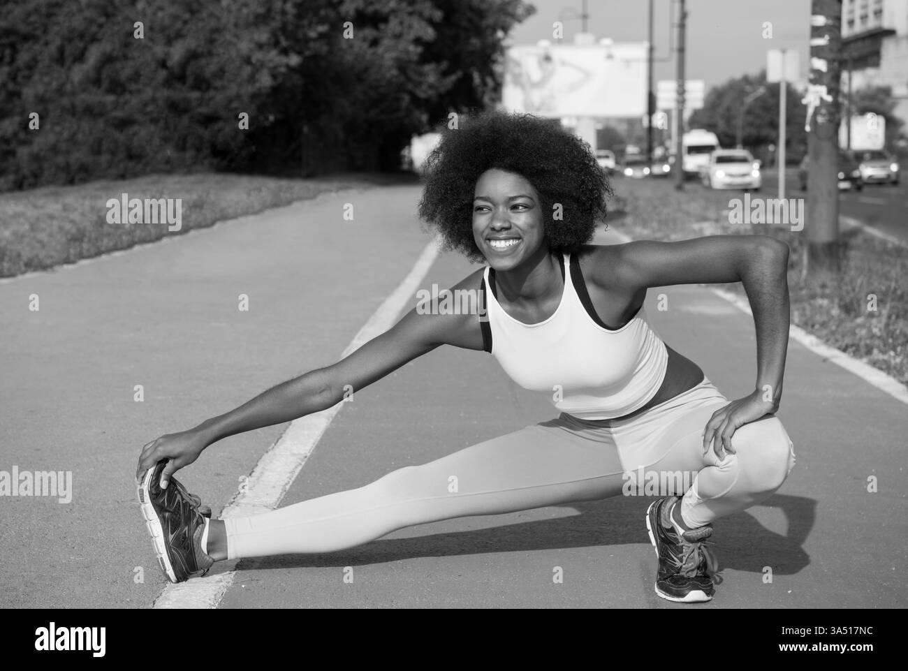 Ritratto di una giovane donna nera che si estende su una strada della città durante una mattinata d'estate soleggiata. Questo momento di fitness all'aperto trasmette fiducia ed energia, rendendolo adatto per l'abbigliamento sportivo, le campagne di benessere e il branding di lifestyle urbano. Foto Stock
