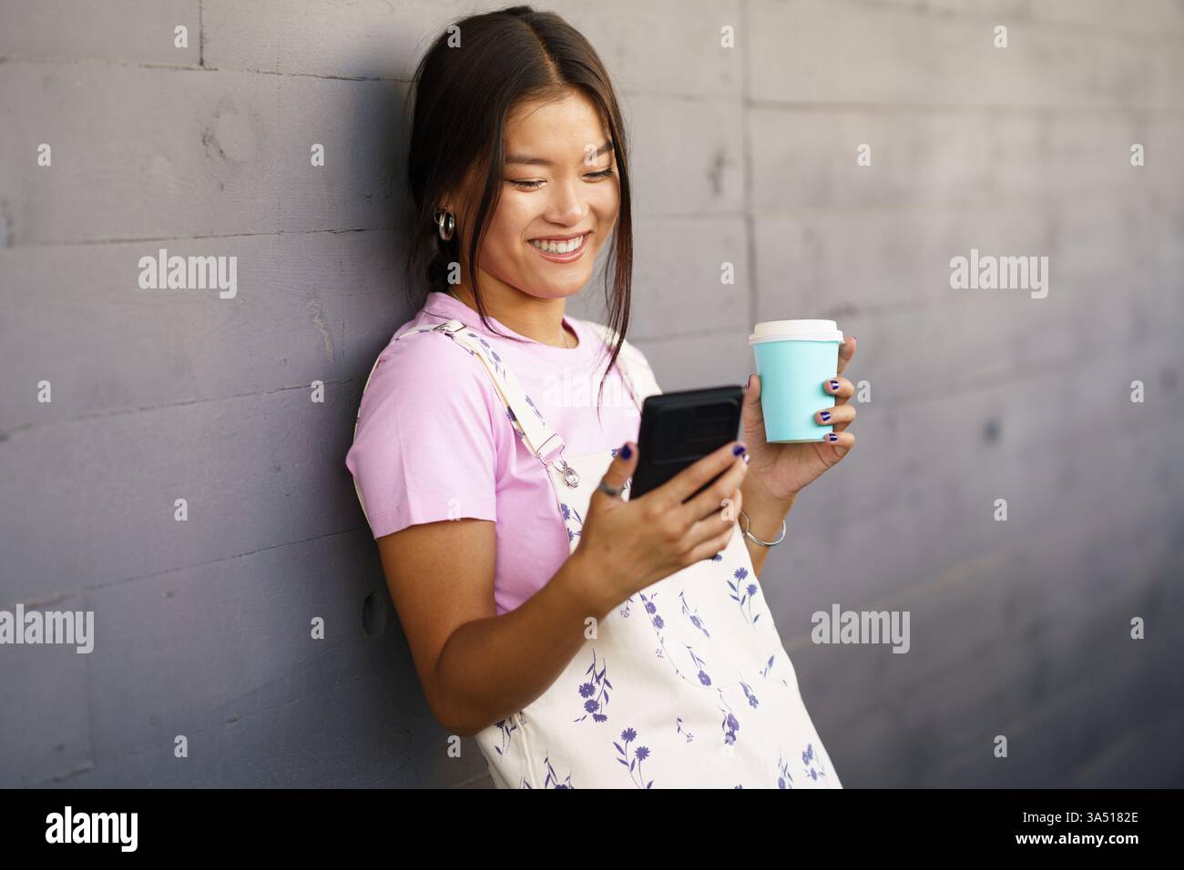 Una donna asiatica ottimista in un abito elegante si gode un caffè da asporto mentre guarda un video sul suo smartphone contro un muro grigio in una strada cittadina. Questo scatto luminoso alla luce del giorno mette in risalto la moda urbana, la navigazione mobile e i momenti sui social media. Ideale per marchi lifestyle, campagne Street style e vita cittadina contemporanea. Foto Stock