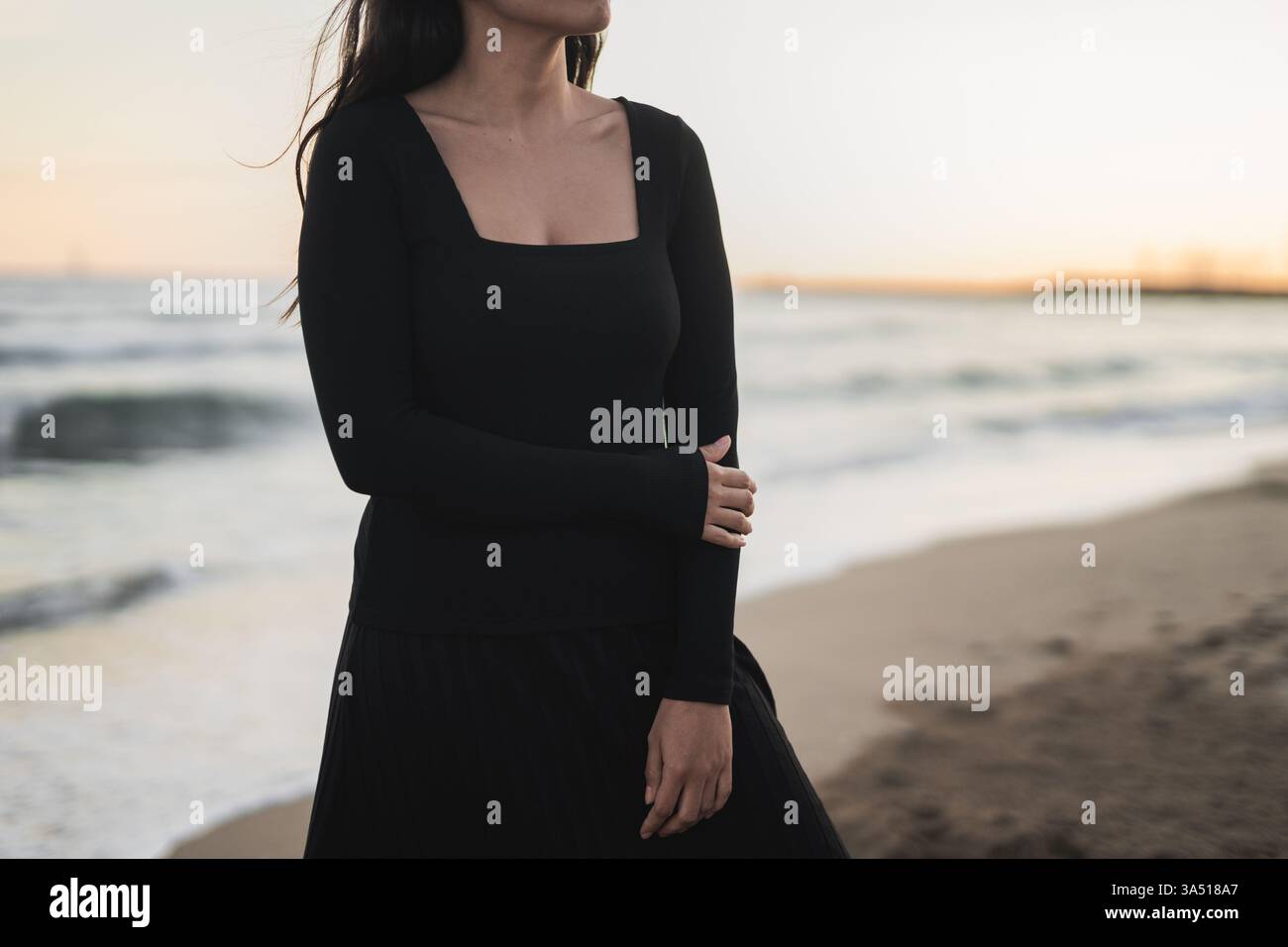 Donna multirazziale con lunghi capelli scuri che indossa un maxi abito nero, si erge su una spiaggia sabbiosa al tramonto, con la mano appoggiata sul braccio. Ottimo per viaggi, vacanze e immagini di moda da spiaggia con un'atmosfera costiera. Foto Stock