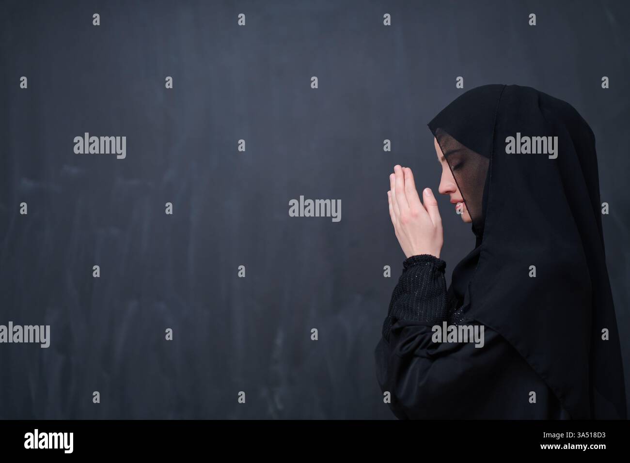 Ritratto di una donna musulmana in un hijab che indossa un abito tradizionale, facendo un gesto di preghiera su uno sfondo scuro. Questa immagine supporta la cultura islamica, la fede e i temi dello stile di vita con un umore calmo e riverente. Foto Stock