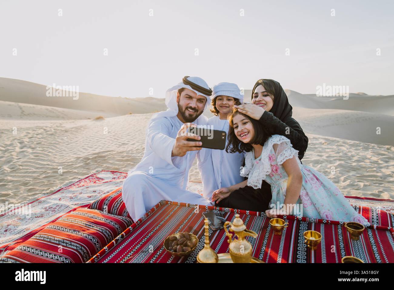 Il padre arabo sorridente fa un selfie con la sua famiglia a un tavolo nel deserto durante il giorno. Questa calda scena di stile di vita familiare si adatta perfettamente alla cultura mediorientale, ai viaggi e alla narrazione del patrimonio culturale. Foto Stock