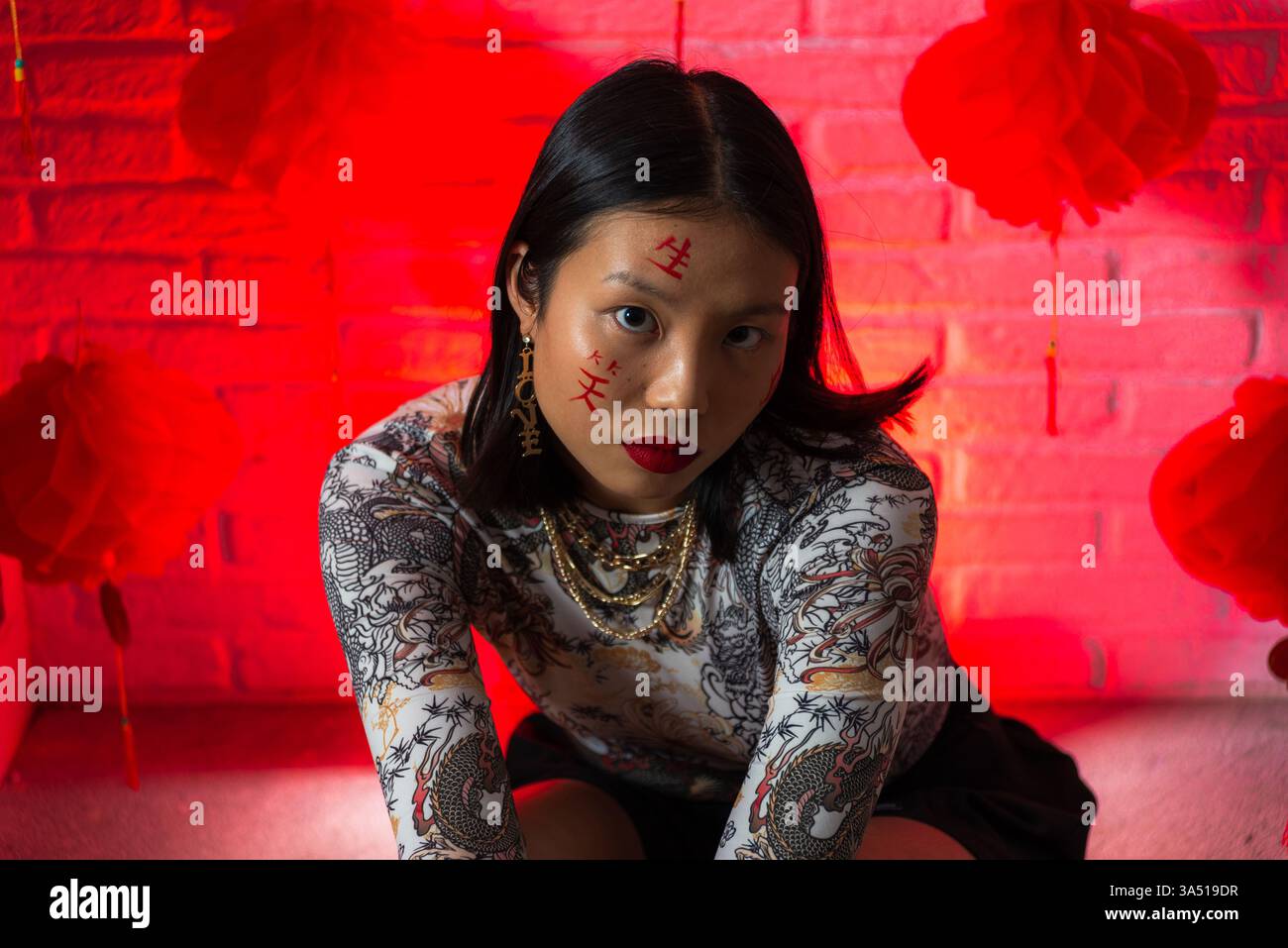 Modella asiatica con pittura facciale seduta su un pavimento di studio, inclinata in avanti per un ritratto audace. L'immagine mostra il trucco artistico e lo Street style contemporaneo, ideale per editoriali di moda. Foto Stock