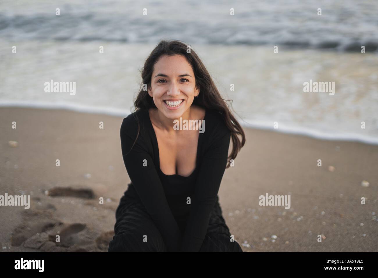 Allegra donna mista in maxi abito nero si siede sulla sabbia in spiaggia durante le vacanze. La posa rilassata e il sorriso soleggiato evocano momenti spensierati sul mare. Foto Stock