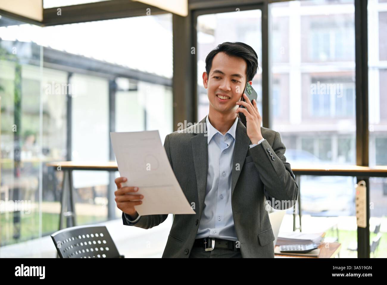 Un uomo d'affari asiatico professionista in tuta formale esamina un rapporto mentre parla su uno smartphone. Un'immagine pronta per l'uso in ufficio o in azienda per argomenti di finanza, consulenza e comunicazione con i clienti. Da utilizzare per aggiornamenti aziendali, riunioni aziendali o contesti telefonici mobili. Foto Stock