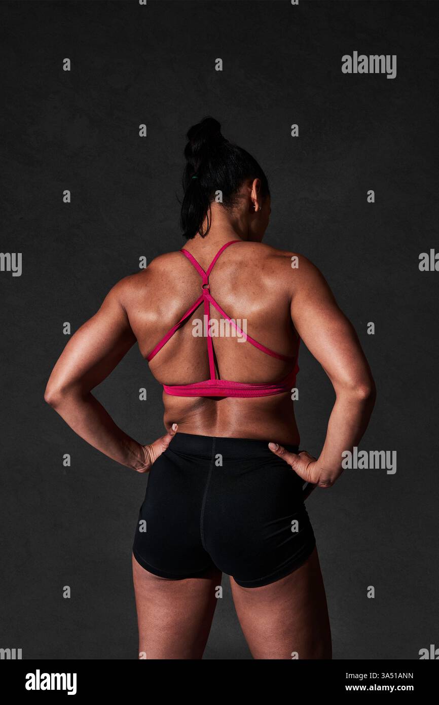 Vista posteriore di un'atleta forte e senza volto con un top rosso e pantaloncini corti sullo sfondo nero. L'immagine enfatizza il fitness, la potenza e un'atmosfera di allenamento stimolante per le campagne di abbigliamento sportivo e benessere. Ideale per il branding in palestra e il marketing del benessere. Foto Stock