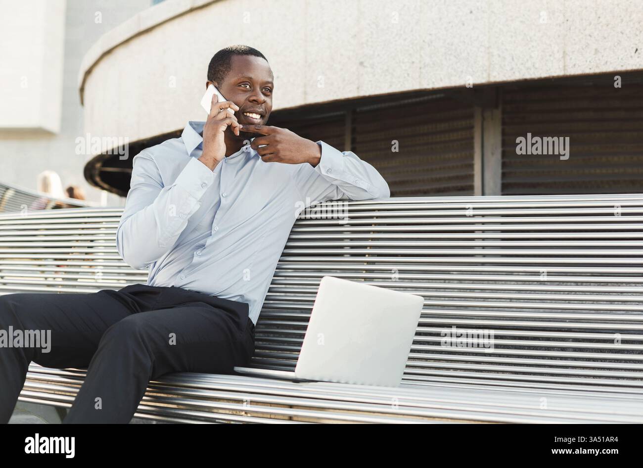 Uomo d'affari Happy Black all'aperto, che utilizza un computer portatile e uno smartphone. L'immagine mostra un agente assicurativo in un abbigliamento casual per affari che si gode un lavoro produttivo all'aperto. Utile per il lavoro mobile, le assicurazioni e le scene aziendali all'aperto. Foto Stock