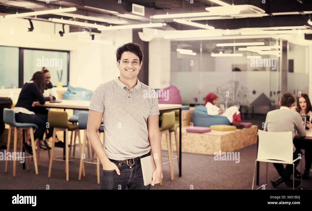 Un uomo d'affari sorridente che tiene in mano un laptop, in tasca con la mano accanto ai colleghi in un ufficio affollato. Questo ritratto aziendale trasmette leadership, lavoro di squadra e un'atmosfera moderna del luogo di lavoro. Foto Stock