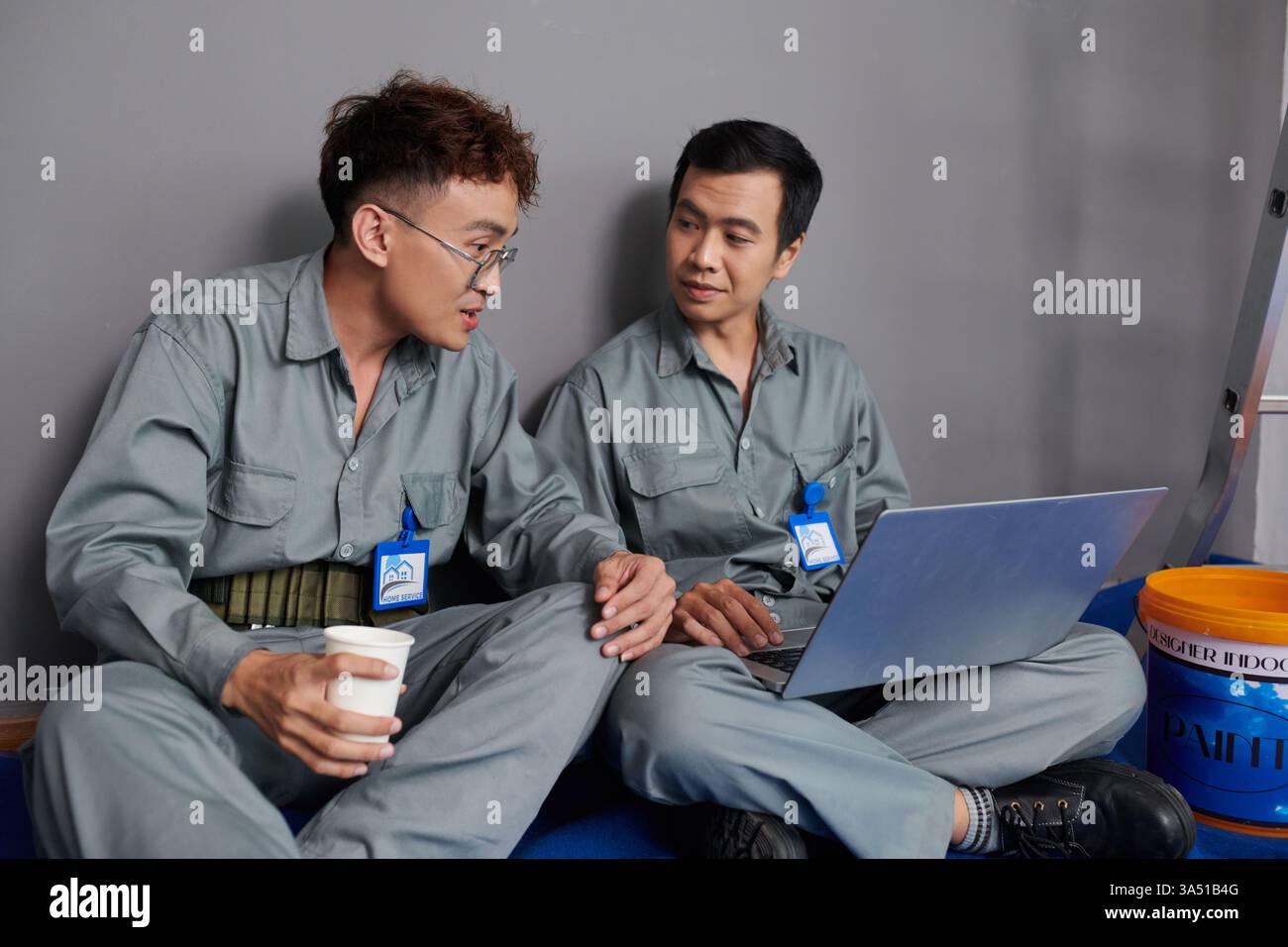 Gli addetti ai servizi domestici siedono sul pavimento con un progetto e un computer portatile, pianificando una ristrutturazione della casa con un caffè. Questa compatta scena di squadra è ideale per la costruzione, il miglioramento della casa o le comunicazioni con i fornitori. Foto Stock