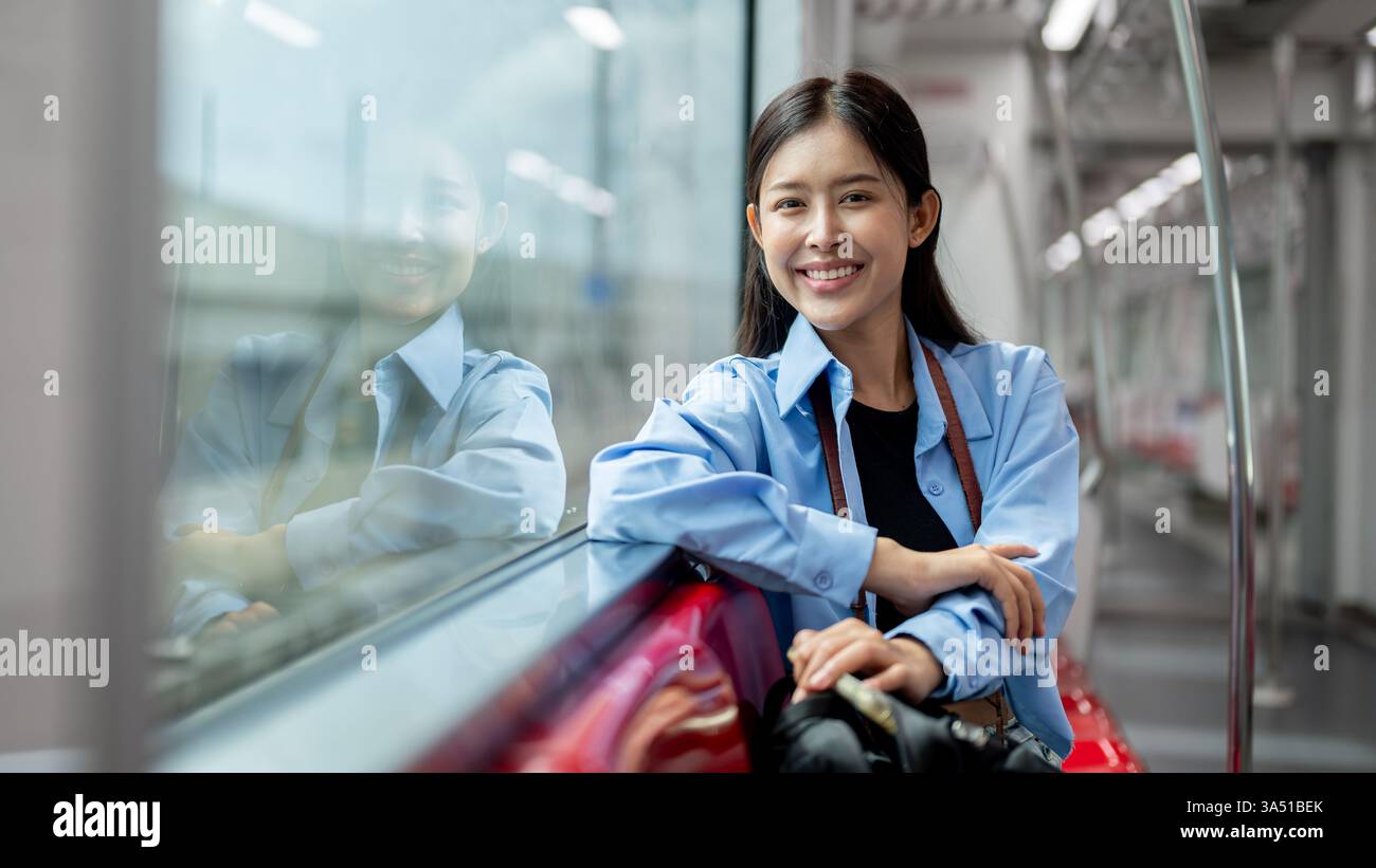 Attraente donna asiatica siede su un treno sopraelevato con il suo zaino, sorridendo alla telecamera. È una pendolare urbana che si gode il viaggio in città in transito come BTS o metropolitana. Ideale per la vita in città, lo stile di vita urbano e le immagini dei trasporti pubblici. Foto Stock