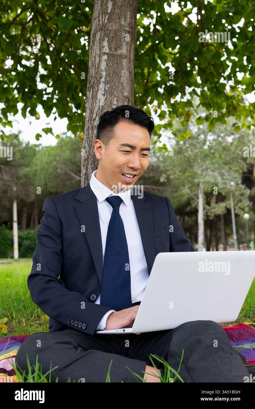 Uomo asiatico sorridente che lavora su un computer portatile mentre è seduto sull'erba in un parco. Lavoro all'aperto e stili di vita flessibili, combinando natura e connettività digitale. Perfetto per lavori in remoto, coworking di parchi e contenuti aziendali mobili. Foto Stock