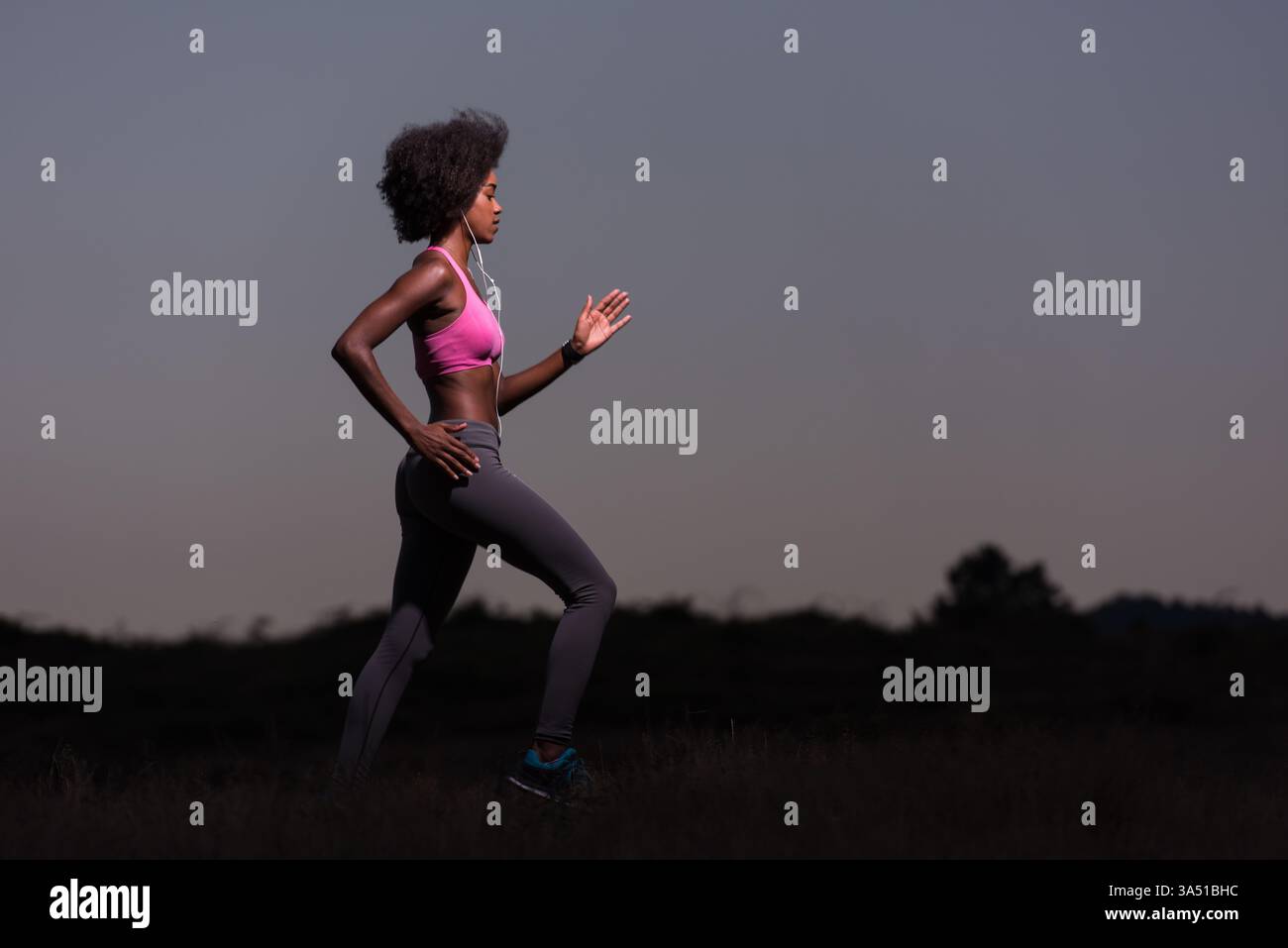 Giovane donna afro-americana che fa jogging all'aperto nella natura durante una bella notte d'estate. Lo shot fitness cattura uno stile di vita attivo, vibrazioni all'aria aperta e branding benessere. Ideale per la salute, l'abbigliamento fitness e le campagne sportive. Foto Stock
