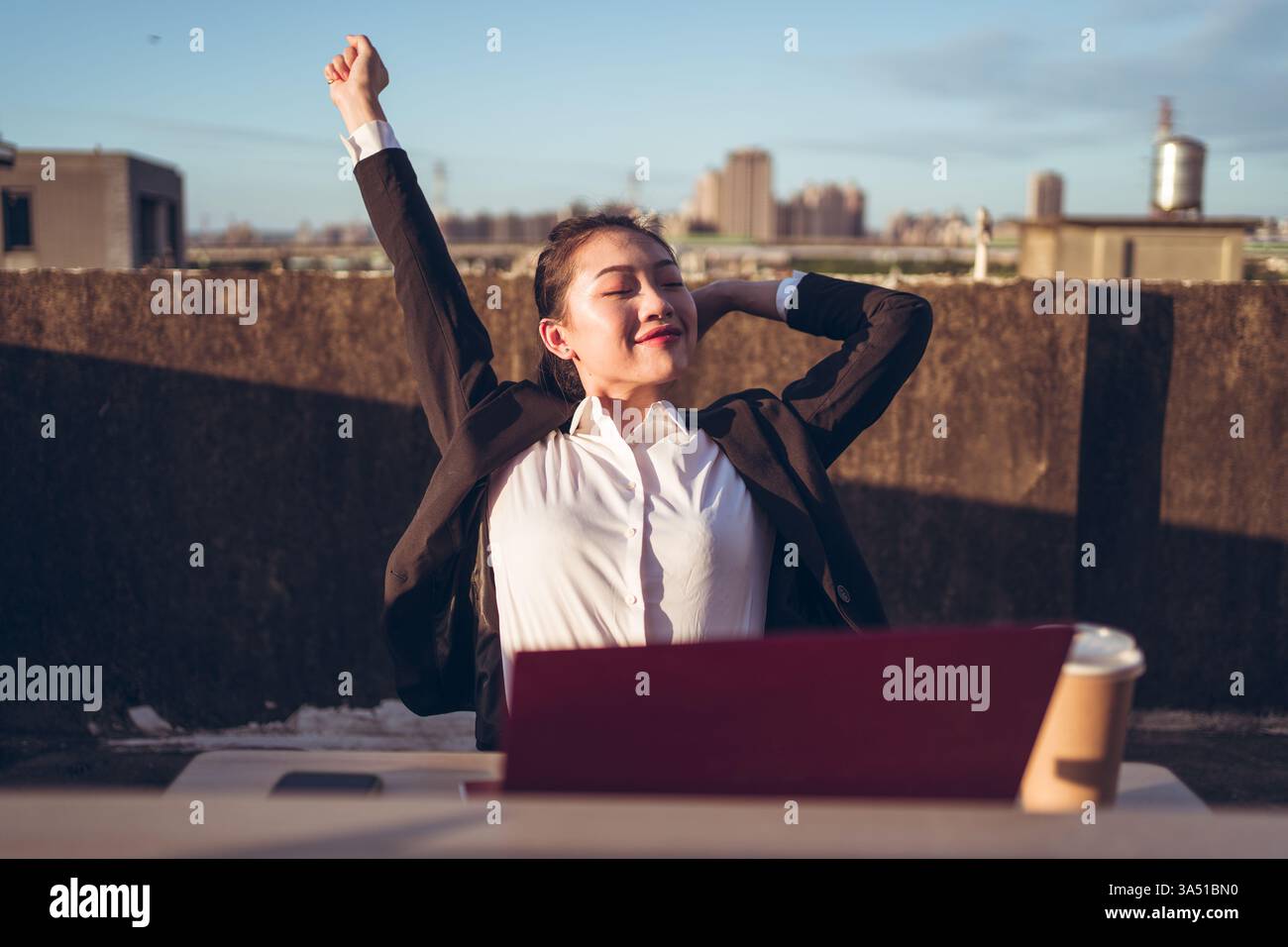 Una donna asiatica siede su un tavolo sul tetto con un portatile, allungando le mani. Questa moderna scena di lavoro a distanza si adatta a storie di lavoro flessibile e stile di vita urbano. Perfetto per la tecnologia, l'imprenditorialità o la grafica per il benessere sul luogo di lavoro. Foto Stock