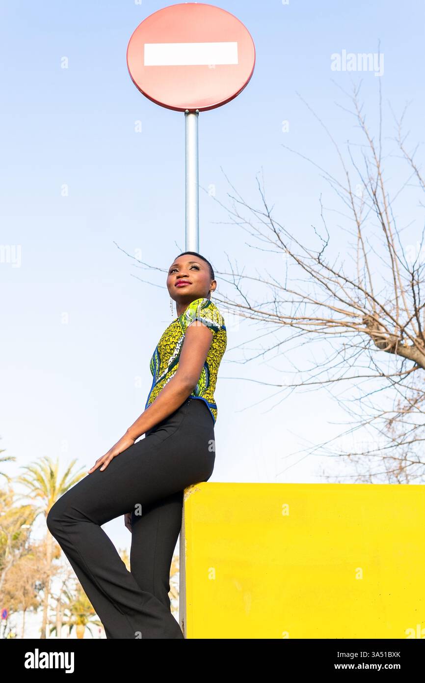 Da una prospettiva bassa, una donna nera con i capelli corti si appoggia su un cartello di divieto di ingresso contro un cielo blu brillante. Lo scatto urbano all'aperto enfatizza l'individualità e la vita di strada. Ottimo per la sicurezza, la segnaletica urbana e le scene estive della città. Foto Stock