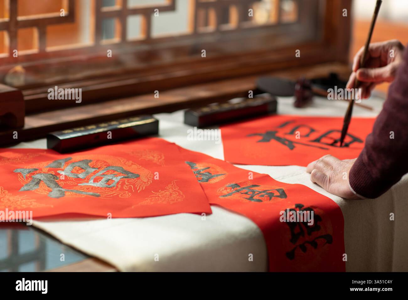 L'uomo cinese anziano scrive calligrafia su distici a casa, catturando un momento festoso di Capodanno. La scena interna enfatizza la cultura tradizionale e la scrittura cinese con decorazioni rosse. Un ritratto ravvicinato e rispettoso di un adulto anziano che ama l'arte della scrittura. Foto Stock