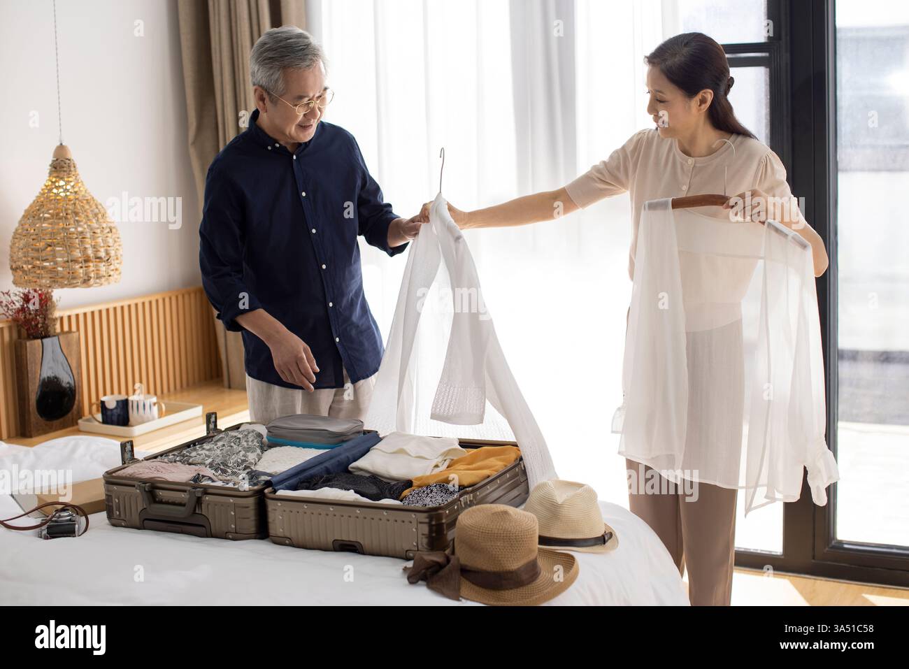 Felice coppia cinese matura che prepara una valigia per le vacanze in una camera da letto d'albergo. Sorridono mentre preparano l'essenziale da viaggio e i vestiti, godendosi insieme. Questa scena di viaggi e stile di vita è ideale per la pianificazione delle vacanze, l'imballaggio e le campagne incentrate su famiglia o anziani. Foto Stock