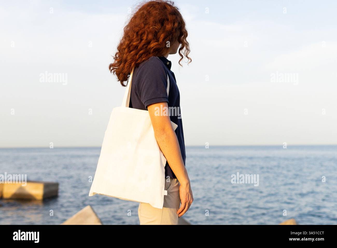 L'uomo ispanico con i capelli ricci che trasportano una borsa si erge sulla spiaggia al tramonto, guardando lontano con ampio spazio per le copie. La luce calda crea un'atmosfera rilassata e pronta per i viaggi. Ideale per la spiaggia, i viaggi e la fotografia di stile di vita. Foto Stock