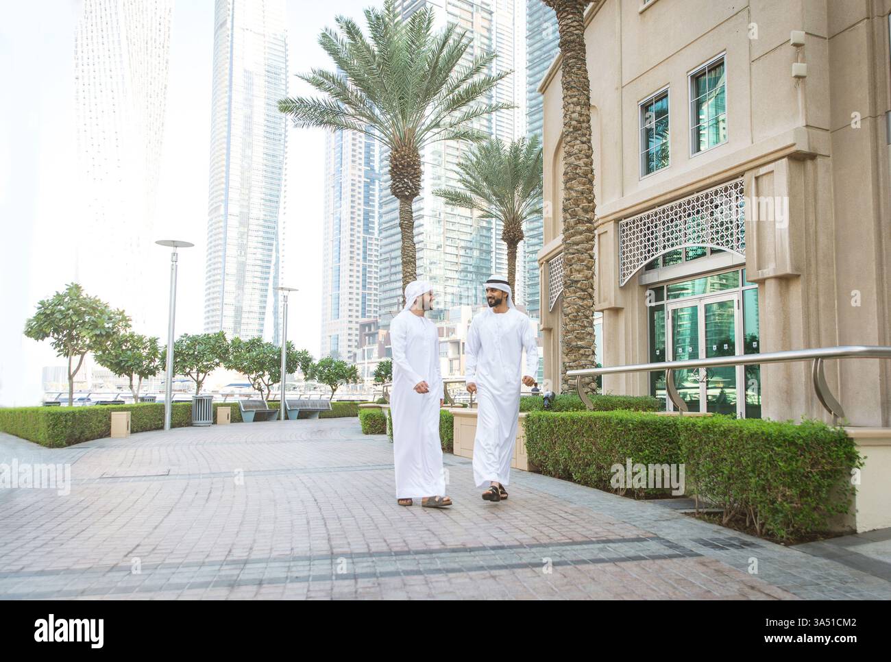 Uomini sorridenti del Medio Oriente in kandura che conversano mentre camminano insieme per le strade della città contro gli edifici nelle giornate di sole Foto Stock