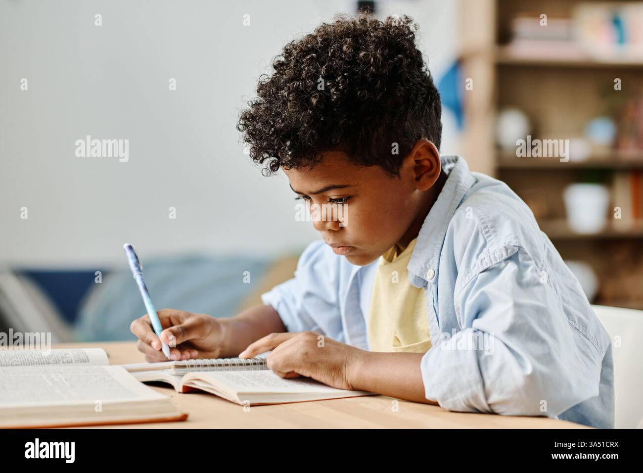 Lo scolaro africano si siede alla sua scrivania a casa, facendo i compiti e prendendo appunti in un taccuino. L'immagine cattura l'apprendimento e lo studio in un ambiente domestico, adatto per l'istruzione, la scuola a casa e il blocco dei contenuti visivi. Foto Stock