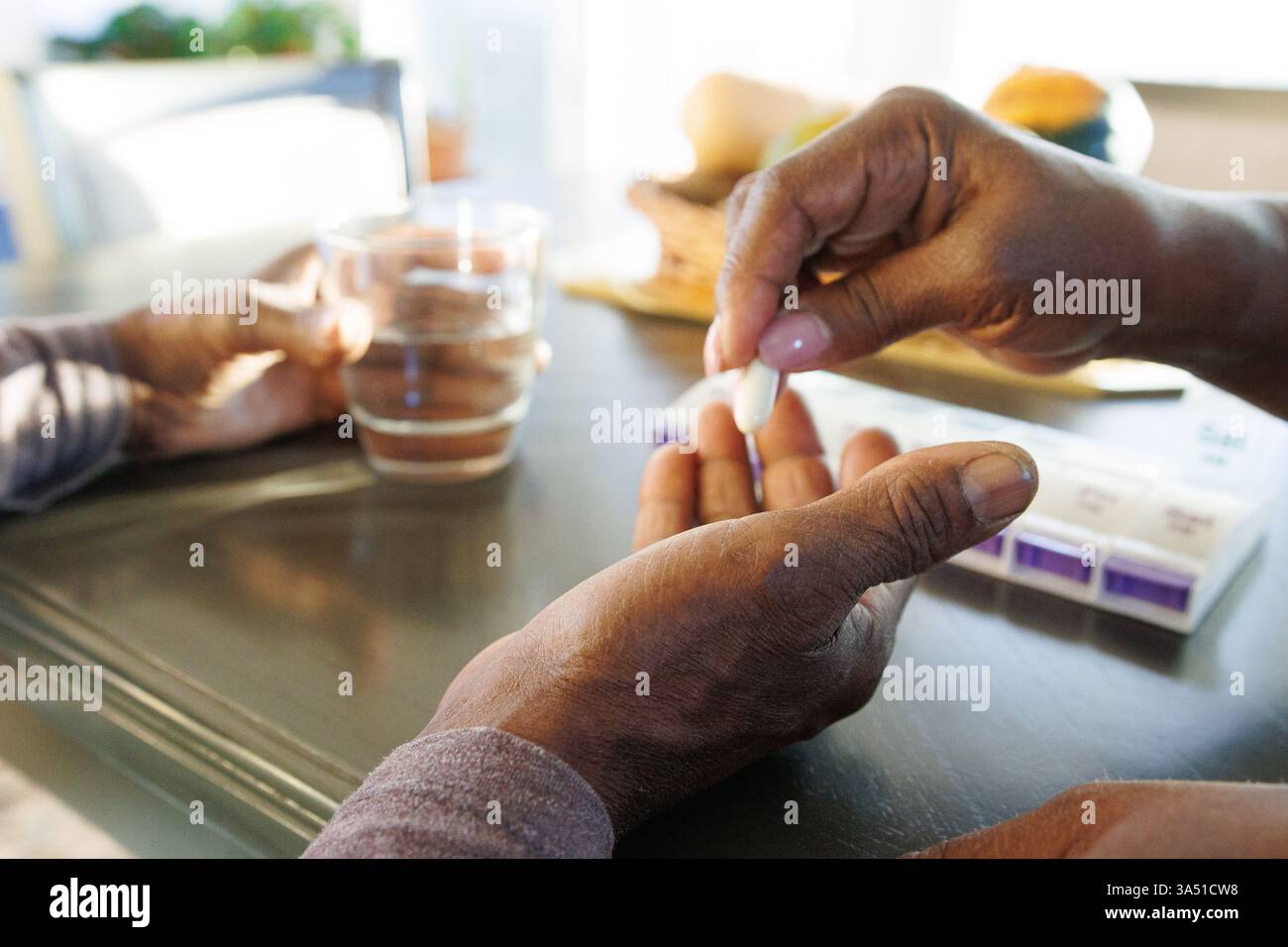 Mani di un operatore sanitario che assiste una persona cara con farmaci a un tavolo da pranzo in un ambiente domestico. Adatto per l'assistenza sanitaria, l'assistenza domiciliare e le immagini del benessere familiare. Trasmette assistenza, supporto e routine quotidiane di salute. Foto Stock