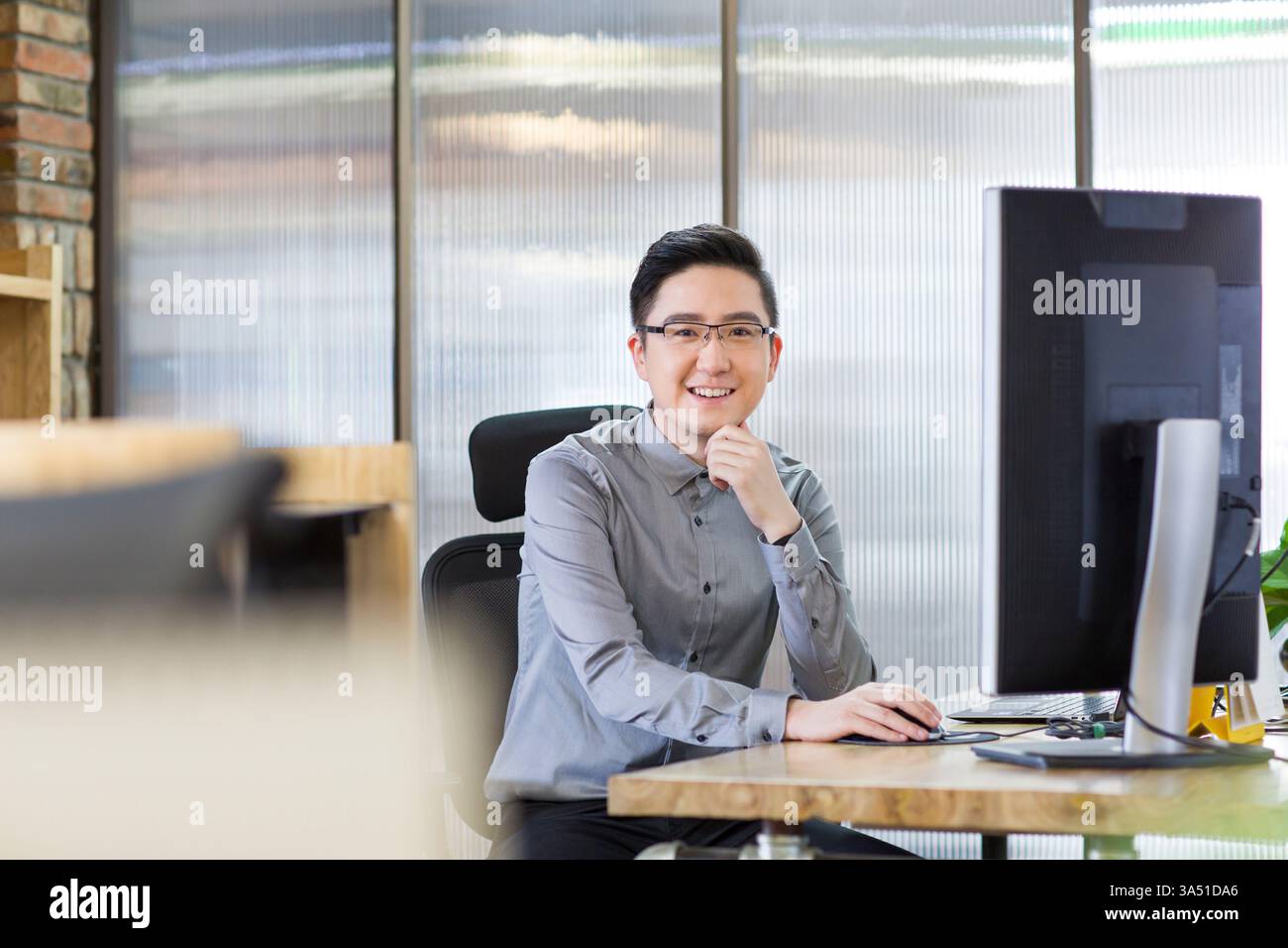 Lavoratore IT cinese seduto al computer in un ufficio moderno. Questa immagine si adatta alle storie dei professionisti tecnologici, dei ruoli IT e dei luoghi di lavoro aziendali. Foto Stock