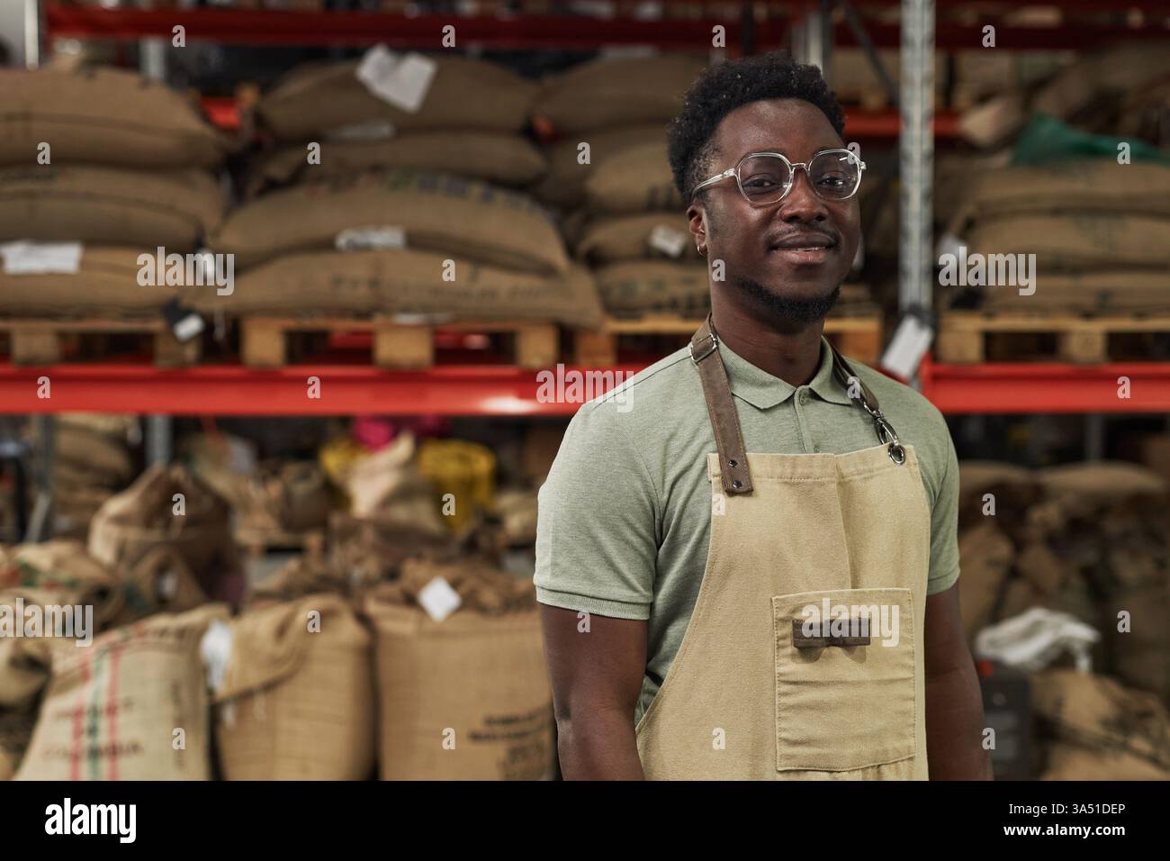 Riprendi il ritratto in vita di un giovane uomo nero che indossa il grembiule e sorride alla macchina fotografica nel magazzino nella torrefazione del caffè, nello spazio per le copie Foto Stock