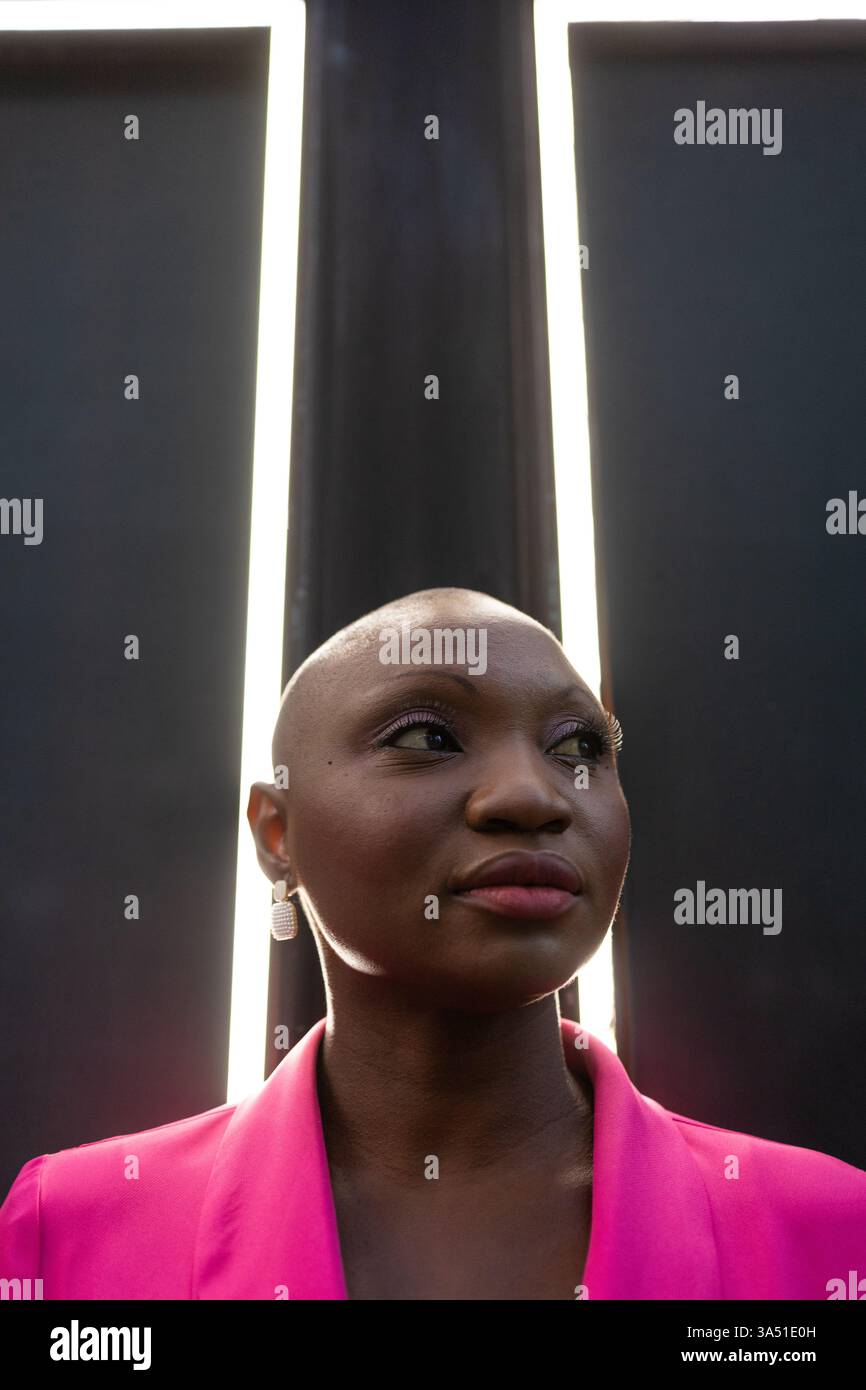 Donna afroamericana sicura di sé con la testa rasata, con un abito rosa e orecchini, si erge davanti a una parete di cornici con luce soffusa che filtra tra tra di loro. La sua espressione e lo stile contemporaneo si adattano alla moda, alla bellezza o alle campagne di lifestyle. Da utilizzare per ritratti editoriali, benessere o branding inclusivo. Foto Stock