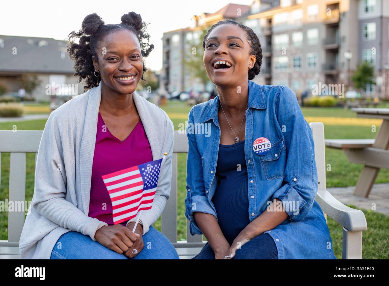 Giovani donne afroamericane siedono su una panchina del parco, ridendo mentre tengono una bandiera americana. Un luminoso momento all'aperto adatto a temi patriottici, impegno civico o eventi della comunità nei parchi pubblici. Cattura l'energia allegra e candida in un parco accessibile con sedia a rotelle. Foto Stock