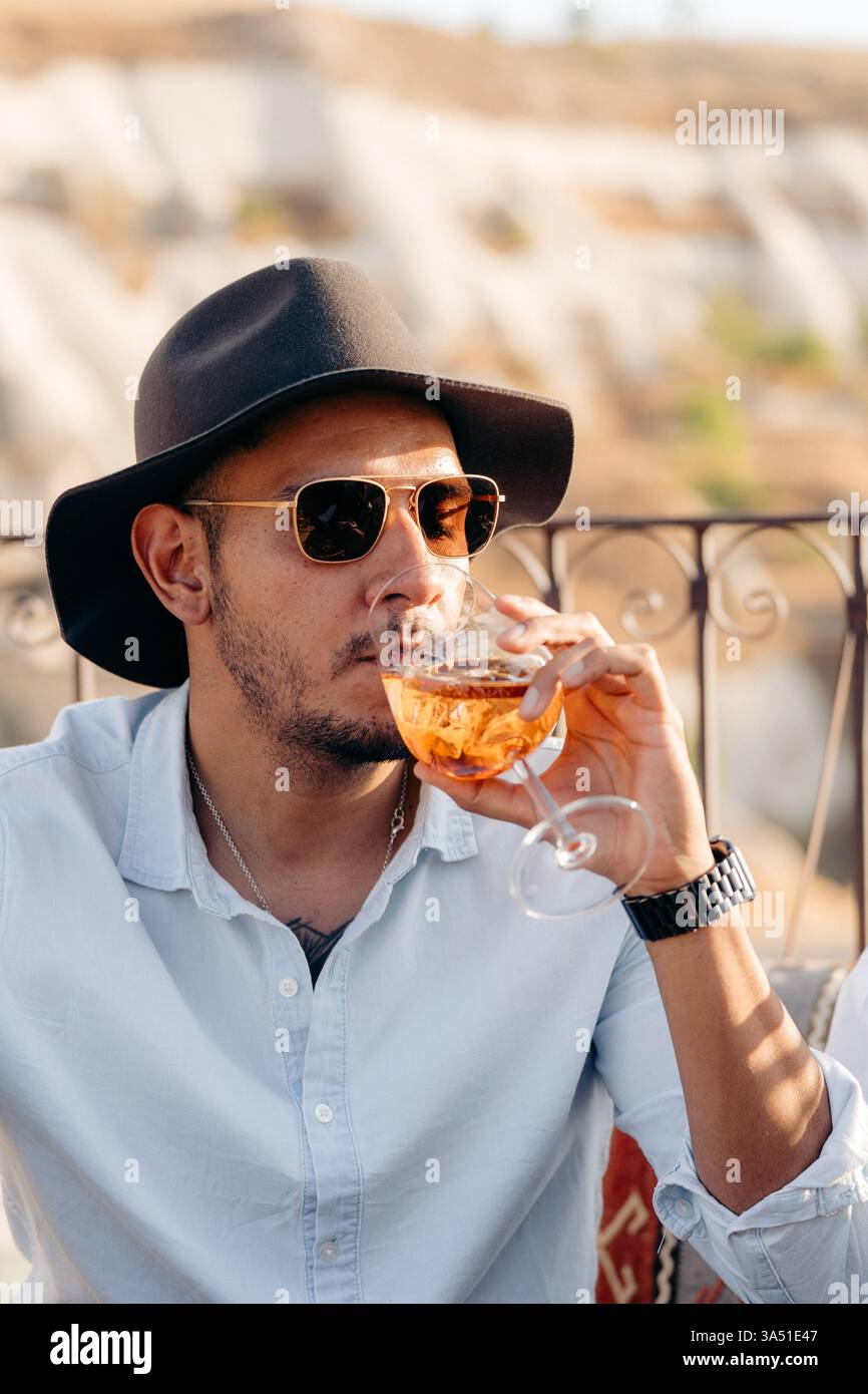 Uomo ispanico che si rilassa su un balcone soleggiato, sorseggiando vino in Cappadocia, Turchia. Questa immagine di stile di vita si adatta ai viaggi, alla vita all'aperto e ai temi del tempo libero diurno. Foto Stock