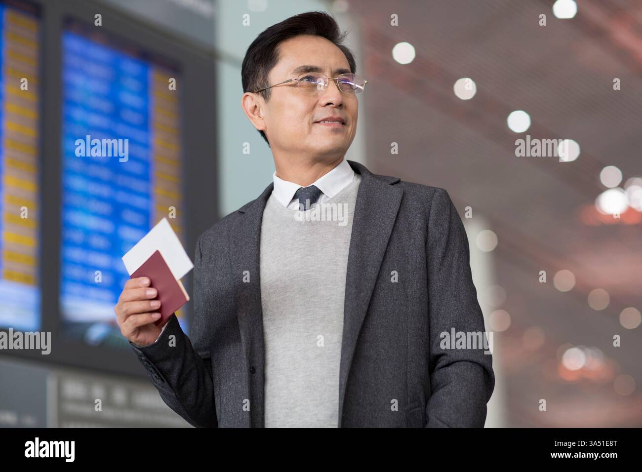 Un uomo d'affari cinese sorridente all'aeroporto, in possesso di un passaporto e di un biglietto aereo. È in piedi con uno sguardo sicuro, pronto per viaggi d'affari o di piacere. Perfetto per viaggi, aeroporti e videografica per la mobilità professionale. Foto Stock