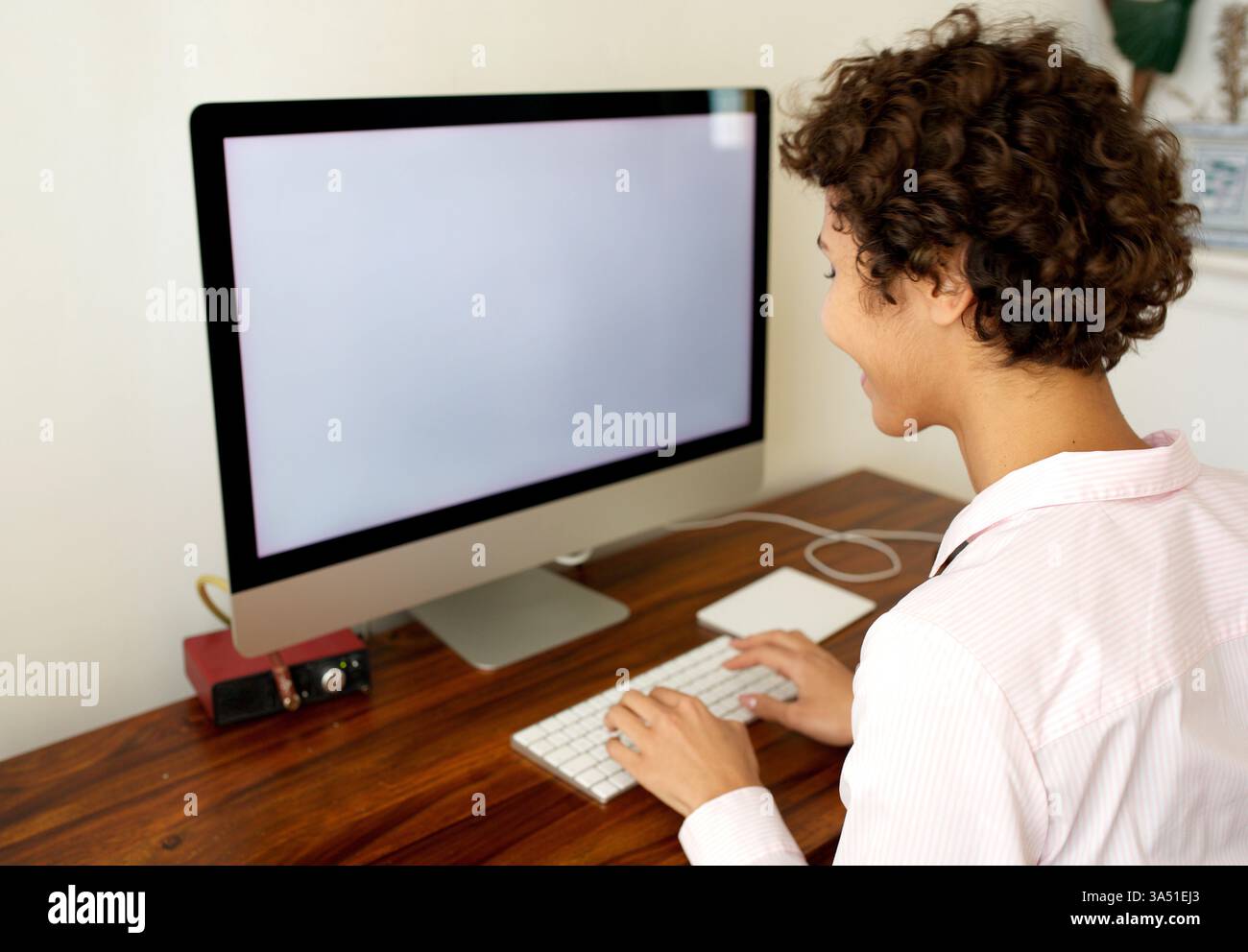 Donna afroamericana sorridente che lavora da casa a una scrivania con un computer. Questa scena supporta argomenti relativi al lavoro da remoto, agli uffici domestici e allo stile di vita digitale. Adatto a contenuti di lavoro e lifestyle con moderne configurazioni di lavoro. Foto Stock