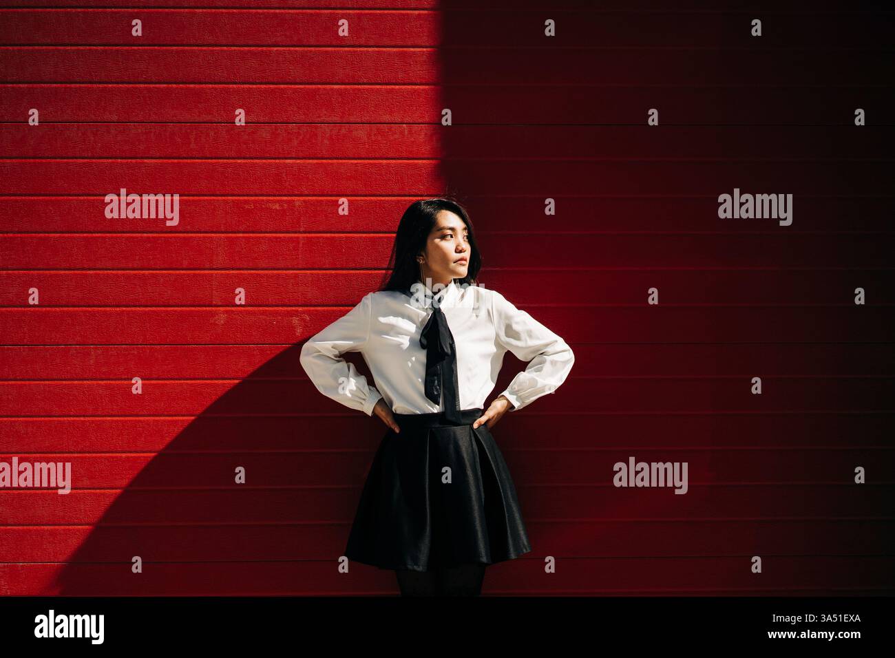 Una donna asiatica con la mano in vita si erge contro un muro rosso, guardando lontano. Questo elegante ritratto urbano è perfetto per le campagne di moda e Street style in contesti contemporanei. Foto Stock