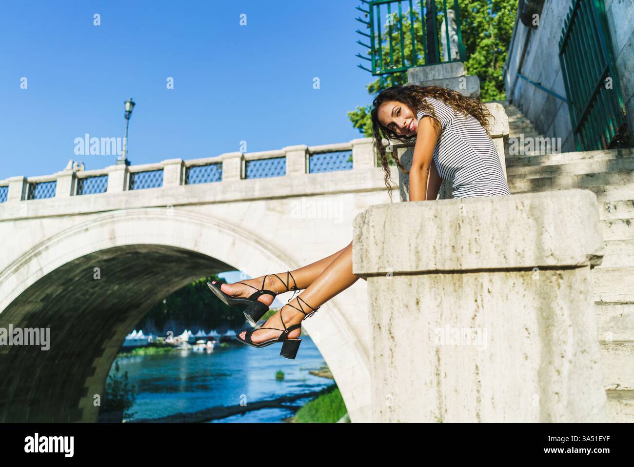 Donna ispanica sorridente siede su cemento vicino a un ponte in un ambiente urbano diurno. La posa premurosa evoca viaggi, esplorazioni e atmosfere informali della città. Ideale per viaggi, turismo e campagne di lifestyle con paesaggi urbani. Foto Stock
