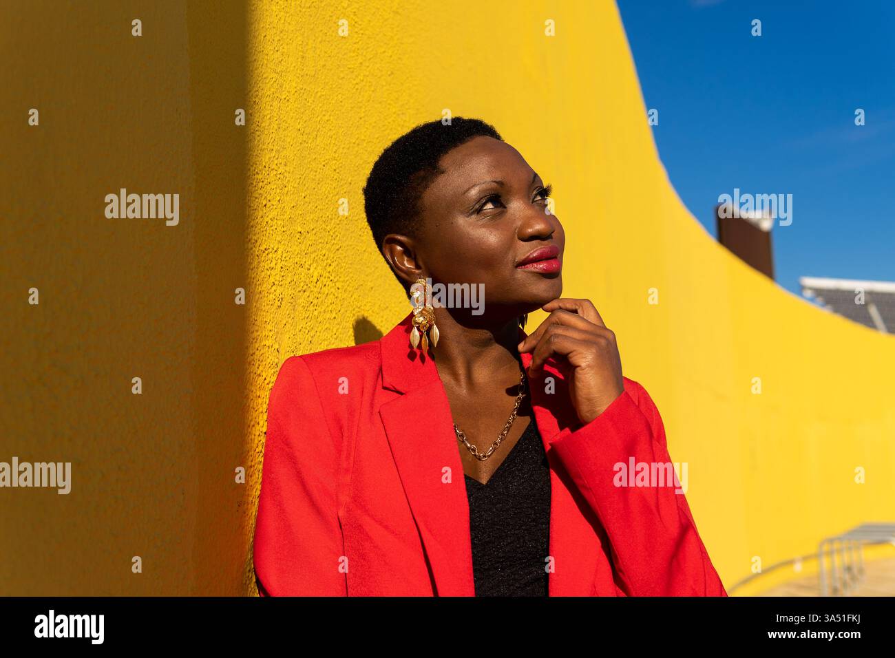 Una donna afroamericana sicura con un abito rosso si erge su una strada soleggiata, appoggiata su un muro giallo. Tocca il mento e si allontana con una posa attenta ed elegante, mettendo in risalto lo stile professionale e la Street fashion. Ideale per la grafica aziendale, di moda e di empowerment femminile in ambienti outdoor. Foto Stock