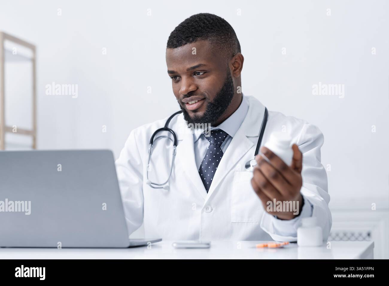 Medico afroamericano sorridente che tiene in mano una bottiglia di pillole mentre lavora al computer portatile in una moderna clinica o in un ufficio. Questa immagine trasmette l'assistenza sanitaria, la professionalità medica e i flussi di lavoro digitali per la salute. Adatto per marketing medico, telemedicina e comunicazioni cliniche. Foto Stock