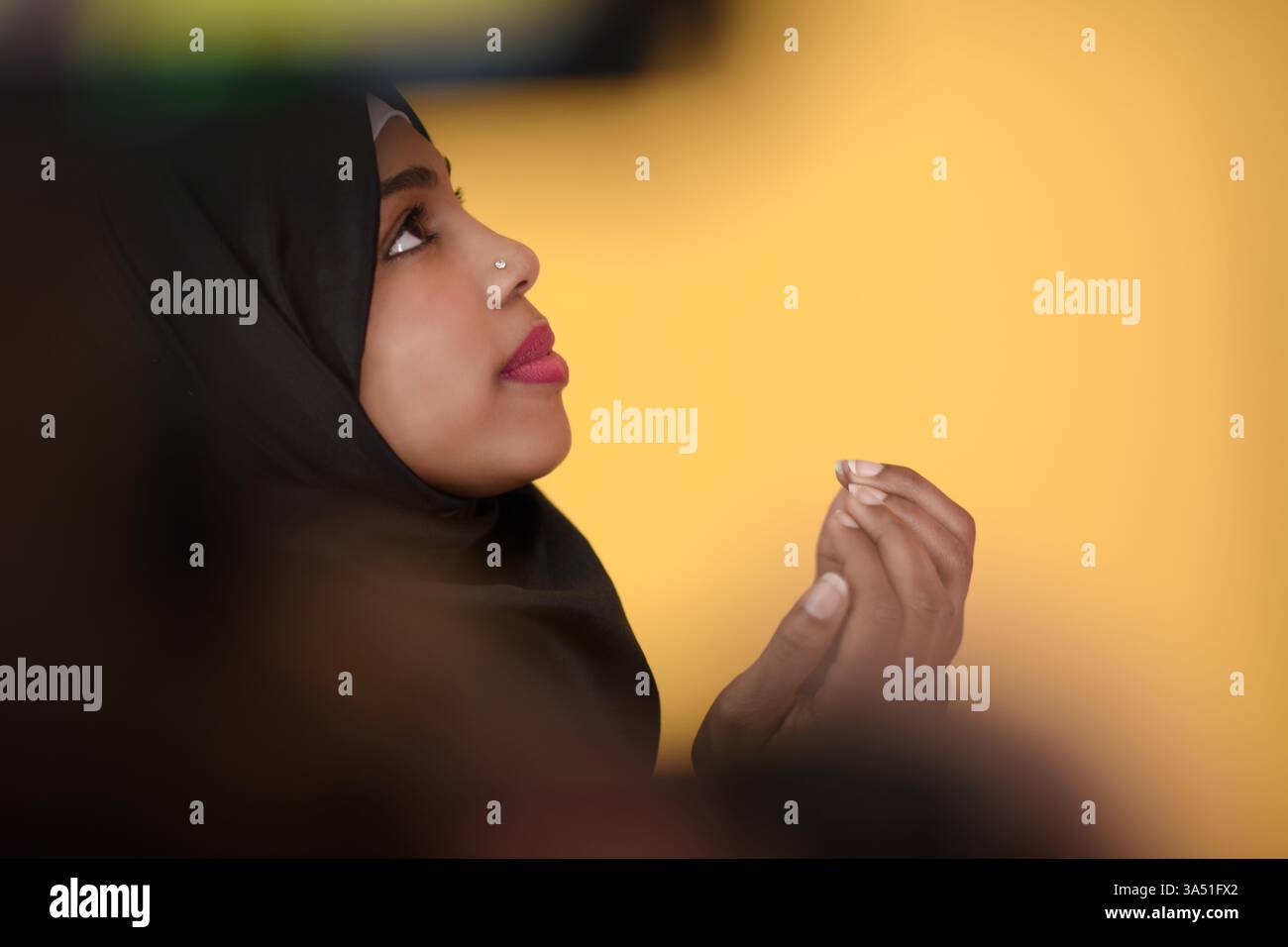 Una seria donna musulmana nera che indossa un hijab guarda verso l'alto con un gesto di preghiera. Lo scatto viene effettuato in uno studio con uno sfondo giallo brillante all'interno. Questa immagine è ideale per temi di fede, preghiera, spiritualità e diversità culturale. Foto Stock