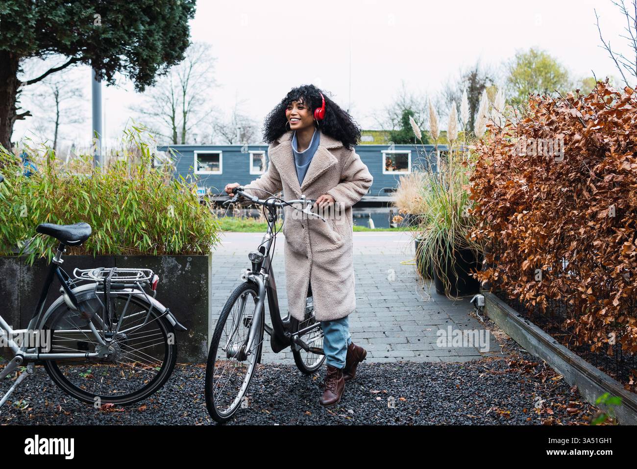 Donna da corsa mista sorridente con capelli ricci che indossano una giacca calda e cuffie che guardano lontano mentre si va in bicicletta per strada Foto Stock Donna da corsa mista sorridente con capelli ricci che indossano una giacca calda e cuffie che guardano lontano mentre si va in bicicletta per strada Foto Stock