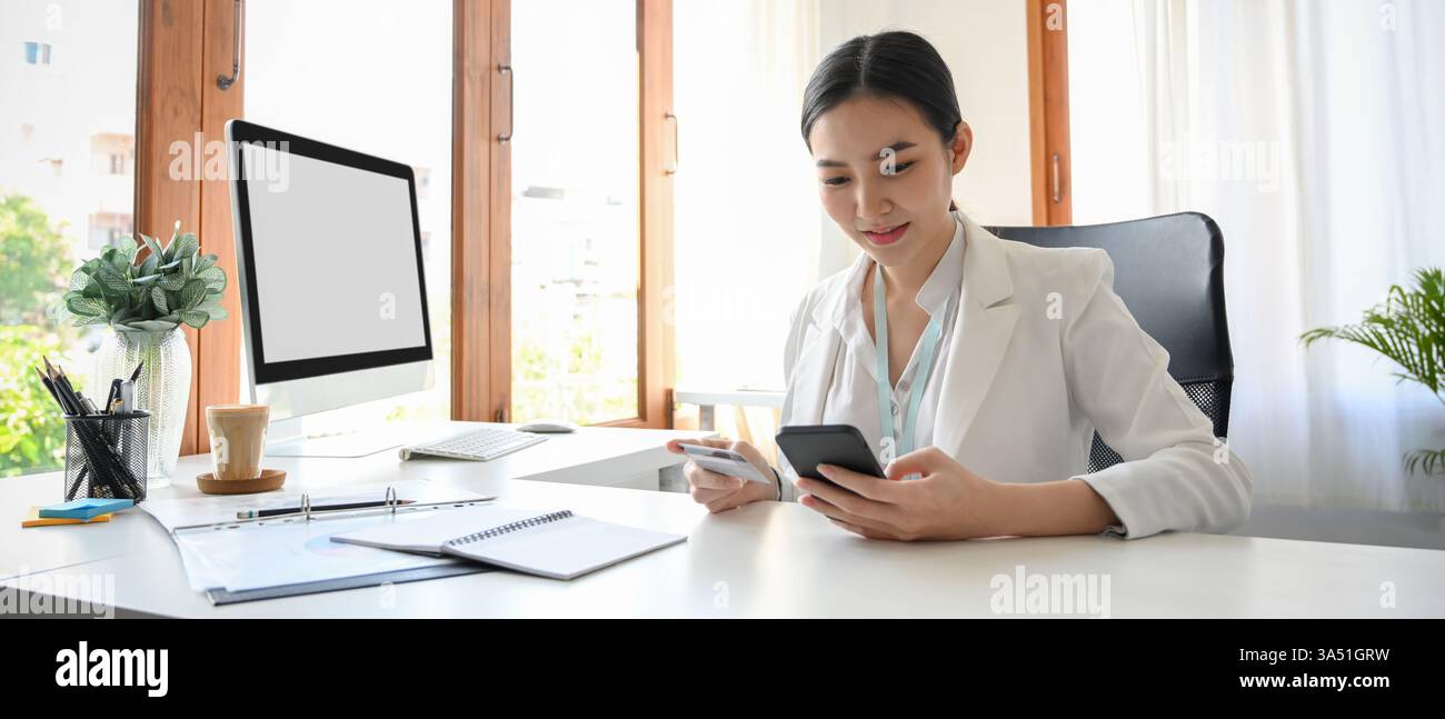 Una donna d'affari asiatica usa una carta di credito per il mobile banking alla sua scrivania. Ideale per il settore bancario, fintech e immagini di lavoro aziendali. Foto Stock