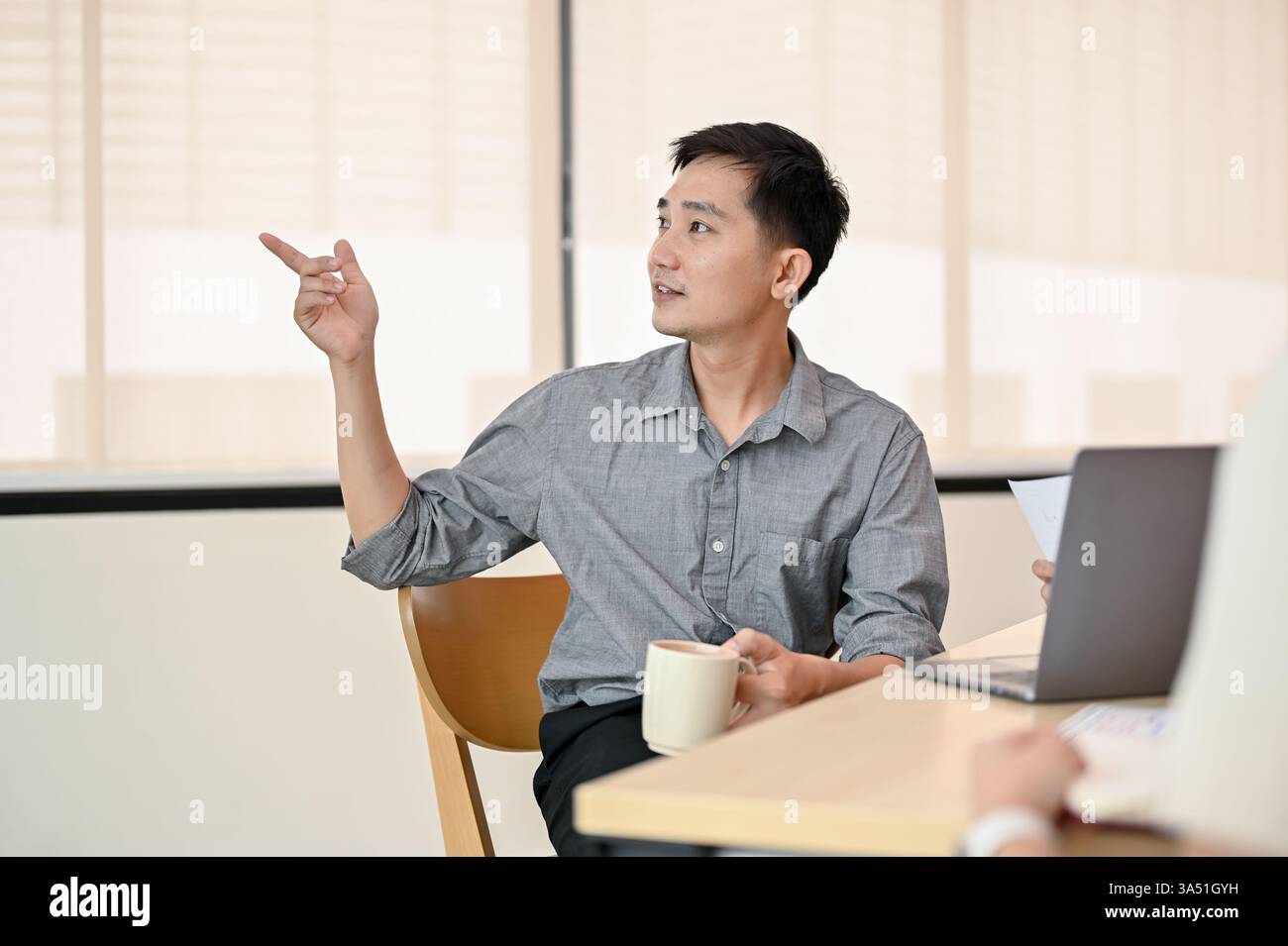 Uomo d'affari asiatico professionista che conduce un incontro con il suo team per sviluppare un nuovo progetto. La scena trasmette il brainstorming, la pianificazione e la progettazione collaborativa in un ambiente aziendale. Adatto per startup tecnologiche, sviluppo di prodotti e discussioni strategiche in Asia. Foto Stock