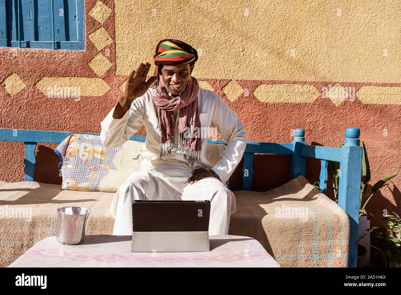 Sincero uomo egiziano in chat video tradizionali con turbante e accappatoio su un tablet mentre si siede su un divano soleggiato sul retro. L'ambiente del cortile fonde cultura e comunicazione moderna. Ideale per lo stile di vita, la tecnologia e la narrazione interculturale. Foto Stock