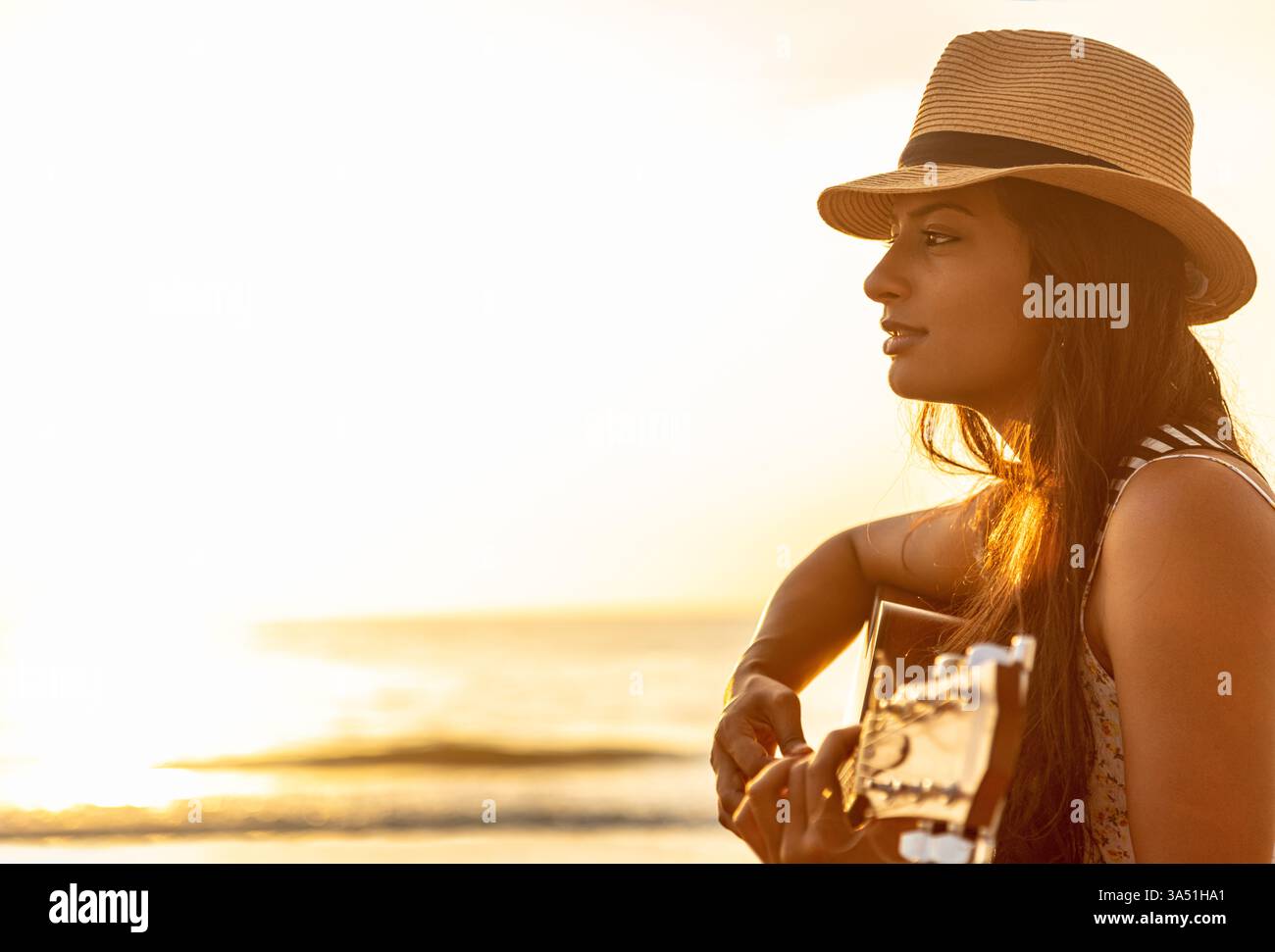 Donna indiana sorridente che indossa un cappello che si rilassa durante le vacanze estive, suona la chitarra sulla spiaggia all'alba. Cattura l'atmosfera rilassata del viaggio e il tempo libero sulla costa. Ideale per viaggi, musica e grafica per le vacanze. Foto Stock