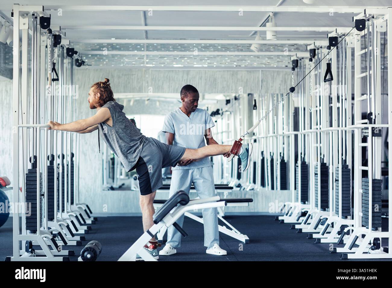 Un terapeuta afroamericano maschile dimostra tecniche di riabilitazione per una gamba ferita utilizzando attrezzature specializzate in un club sportivo. Questa scena sanitaria e fisioterapia enfatizza l'assistenza ortopedica e il recupero delle lesioni in una palestra interna. Perfetto per campagne di medicina sportiva, riabilitazione e servizi di riabilitazione professionale. Foto Stock
