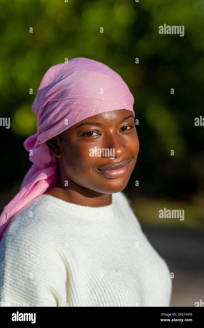 Una donna afroamericana che indossa un velo rosa si erge in un parco alberato, presentando un ritratto calmo e avvicinabile. Questa immagine di stile all'aperto naturale si adatta al benessere, alla cultura e a contesti di moda inclusivi. Ideale per campagne naturalistiche con donne diverse. Foto Stock